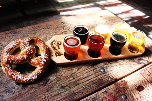 A wooden flight board with eight small beer glasses and a large pretzel on a rustic table.