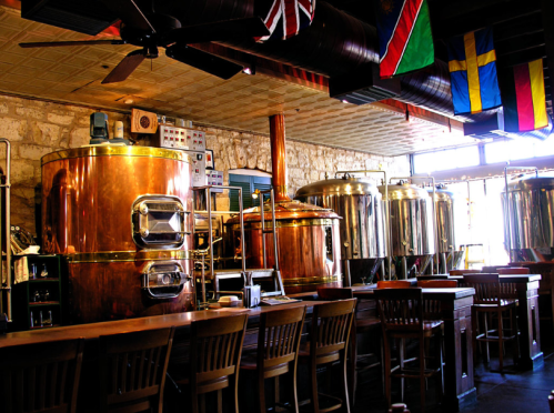 Interior of a brewery featuring large copper and stainless steel brewing tanks, with flags and wooden bar seating.