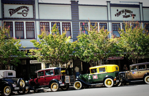 A row of vintage cars parked in front of a brewery and restaurant, surrounded by trees and historic buildings.