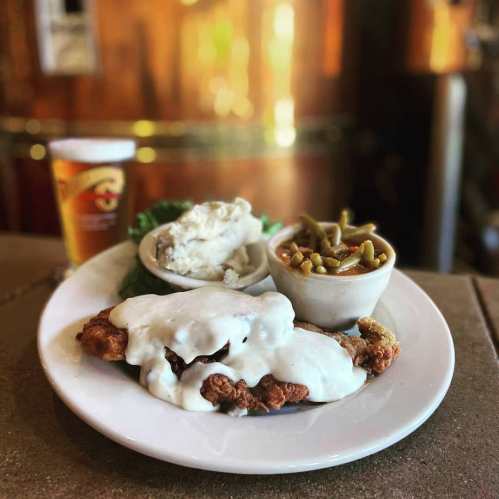 A plate of fried chicken topped with white gravy, served with mashed potatoes, green beans, and a beer on the side.