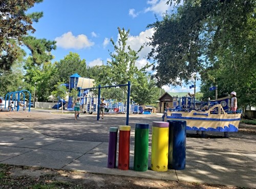 A colorful playground with swings and play structures, surrounded by trees and blue skies.