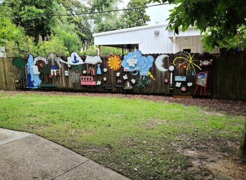 A colorful mural on a wooden fence features various nature and weather-themed decorations in a grassy area.