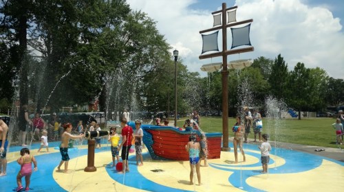 Children play in a splash pad featuring a pirate ship, surrounded by water fountains and a sunny park setting.