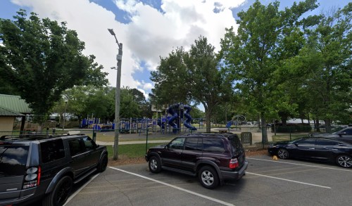 A playground with blue equipment surrounded by trees, viewed from a parking area with parked cars.