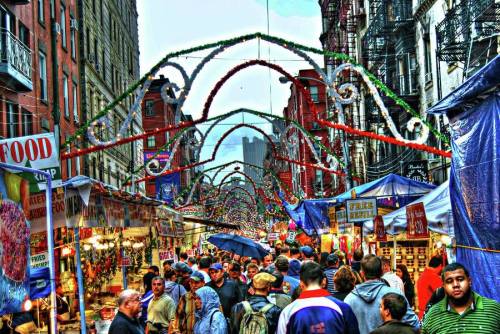 A bustling street market decorated with festive arches, filled with people browsing food stalls and colorful tents.