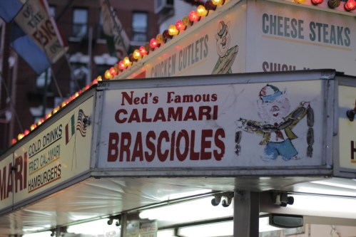 Sign for "Ned's Famous Calamari" and "Brasciole" at a food stand, featuring colorful lights and a cartoon chef.