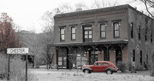 A vintage red car parked in front of a historic brick building with a sign reading "Chester" nearby.