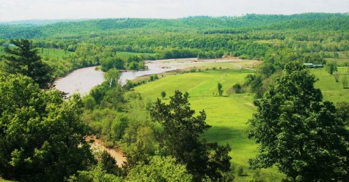 A scenic view of a river winding through lush green fields and trees under a bright blue sky.