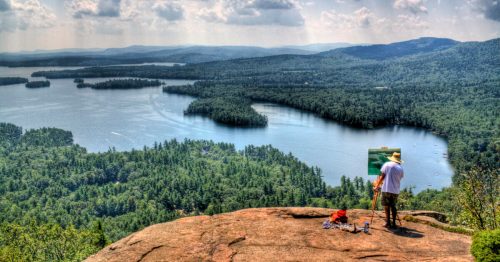 An artist paints a scenic view of a lake surrounded by lush forests and mountains from a rocky overlook.