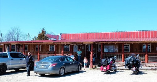 A rustic building with a red roof, parked cars, and people standing outside on a sunny day.