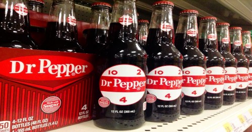 A row of glass bottles of Dr Pepper soda on a store shelf, with a box of four bottles in the foreground.