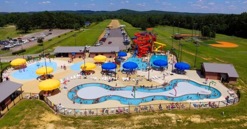 Aerial view of a vibrant water park with pools, slides, and sun umbrellas, surrounded by green fields and parking.