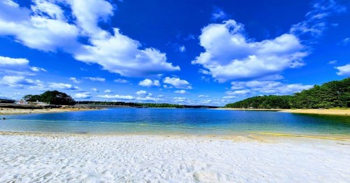 A serene beach scene with clear blue water, white sand, and fluffy clouds under a bright sky.