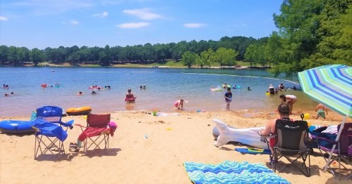 A sandy beach by a lake with people swimming, playing, and relaxing under colorful umbrellas on a sunny day.