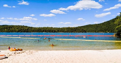 A sandy beach by a lake with swimmers, surrounded by green hills and a blue sky with fluffy clouds.