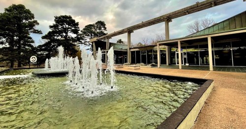 A modern building with large glass windows beside a fountain with water jets, surrounded by trees and cloudy skies.