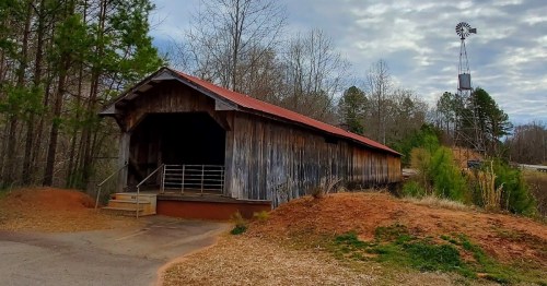 A rustic covered bridge with a red roof, surrounded by trees and a windmill in the background under a cloudy sky.