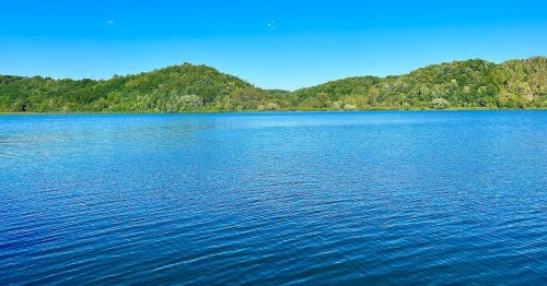 A serene lake surrounded by lush green hills under a clear blue sky. Gentle ripples on the water's surface.