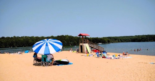 A sandy beach with umbrellas, people relaxing, and a lifeguard tower by a calm lake surrounded by trees.