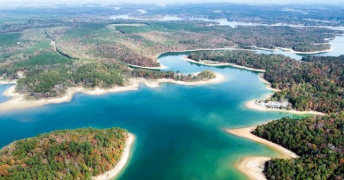 Aerial view of a winding lake surrounded by lush green forests and autumn-colored trees.