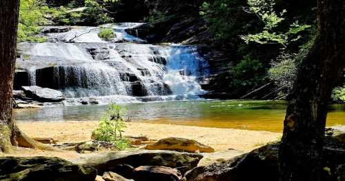 A serene waterfall cascades into a calm pool, surrounded by lush greenery and sandy banks.