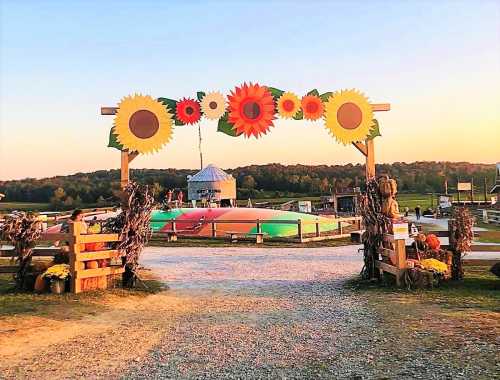 A colorful arch adorned with sunflowers welcomes visitors to a farm at sunset, with a carnival area in the background.