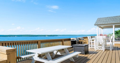 A scenic view of a deck overlooking a calm lake, featuring picnic tables and outdoor seating under a clear blue sky.