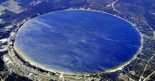 Aerial view of a large, circular lake surrounded by trees and roads, reflecting a clear blue sky.