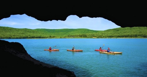 Four kayakers paddle on a calm lake, framed by a cave opening with lush green hills in the background.