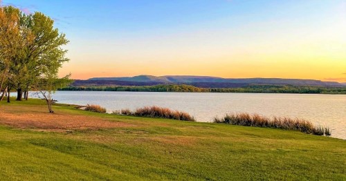 A serene lakeside view at sunset, with grassy shores, trees, and distant hills under a colorful sky.