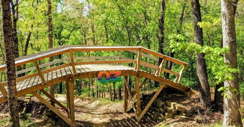 A wooden bridge arches over a dirt path, surrounded by lush green trees in a forested area.