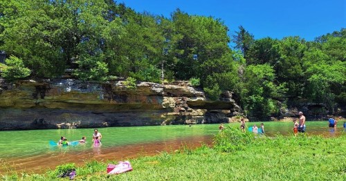 A sunny day at a green river with people swimming and relaxing by the water, surrounded by lush trees and rocky cliffs.
