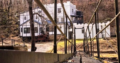 A bridge leads to a white castle-like building surrounded by trees and a hilly landscape.
