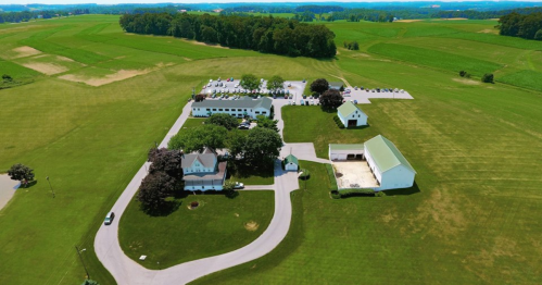 Aerial view of a green landscape featuring a large building complex, parking area, and surrounding fields.