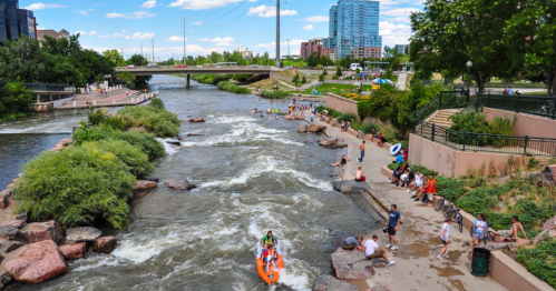 A lively river scene with people kayaking, relaxing on the banks, and enjoying a sunny day in a city park.