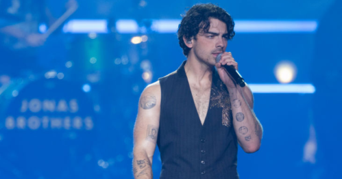 A male singer with tattoos performs on stage, wearing a sleeveless vest, with a band in the background.