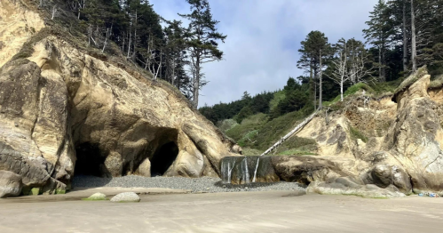 A scenic beach with rocky cliffs, a small waterfall, and trees in the background under a cloudy sky.