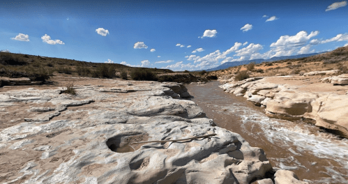 A rocky riverbank with flowing water under a blue sky dotted with clouds. Desert landscape in the background.