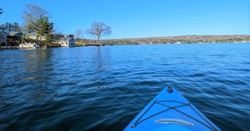 A view from a kayak on calm water, with trees and houses along the shore under a clear blue sky.