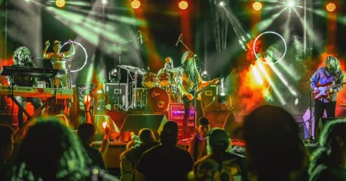 A lively band performs on stage with colorful lights, while an engaged audience watches from below.