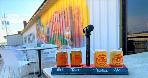 A wooden flight board with four small beer glasses in front of a colorful mural and outdoor seating area.