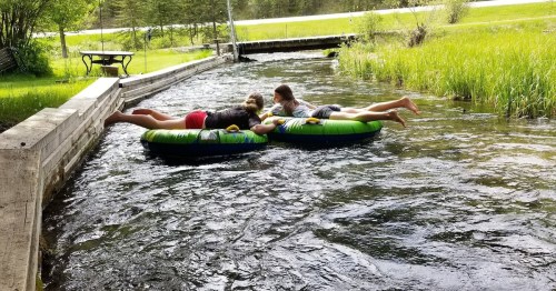 Two children lying on colorful inner tubes in a flowing stream, surrounded by green grass and trees.
