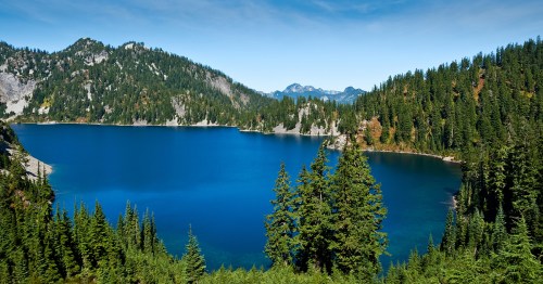 A serene lake surrounded by lush green forests and mountains under a clear blue sky.