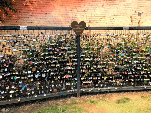 A fence covered in colorful padlocks, symbolizing love, against a brick wall. A heart-shaped sign is visible at the top.