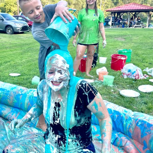 A child pours blue paint over a smiling woman covered in colorful splashes, with buckets and paint containers around them.