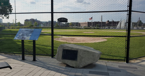 A baseball field behind a fence, with a stone monument and informational plaque in the foreground.