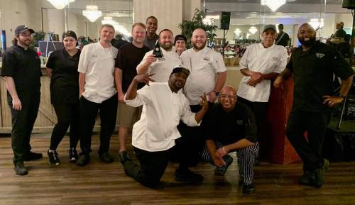 A group of chefs and kitchen staff pose together in a restaurant, smiling and celebrating.