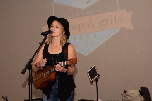 A woman sings into a microphone while playing a ukulele at a music festival, with a logo in the background.