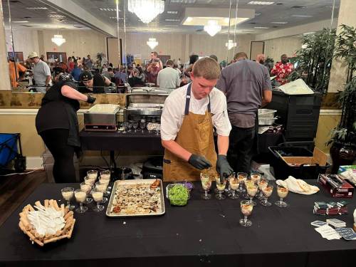 A chef in an apron prepares small dishes at a bustling event with guests in the background.
