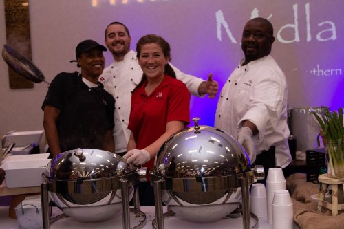 Four chefs smile behind serving trays at a culinary event, with one giving a thumbs up.
